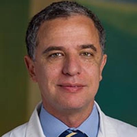 Headshot of Dr. Sadek. He is wearing a shirt, tie, and lab coat in front of a dark green background.