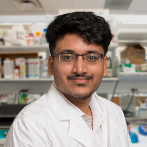 a man with dark hair and mustache wearing a white lab coat standing in a research laboratory