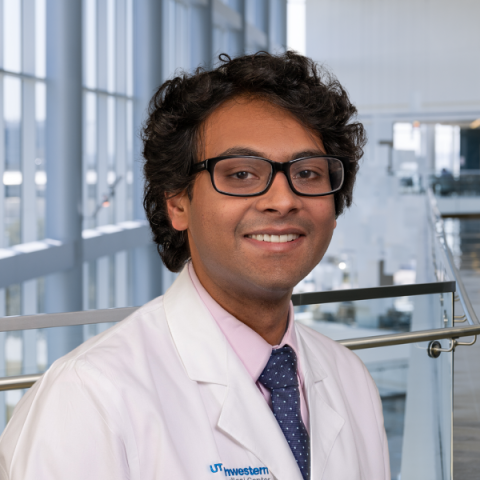 a man with dark curly hair and glasses wearing a white lab coat .and tie standing in a hospital lobby.