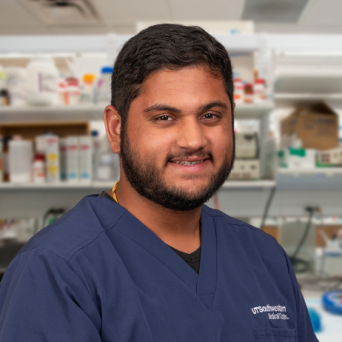 a man with dark hair and beard wearing blue scrubs standing in a research laboratory