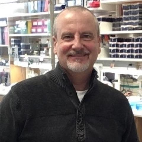 Dr. Farrar smiling, with short cropped gray hair and beard, wearing a black pullover, standing in a lab.