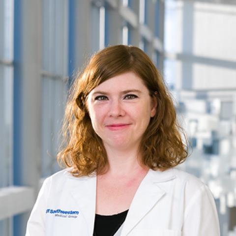 Woman with auburn shoulder-length hair wearing a lab coat, standing in front of large industrial windows.