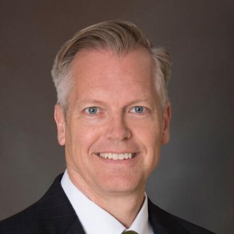 Man with gray hair and blue eyes wearing a black jacket over a white button-down shirt.